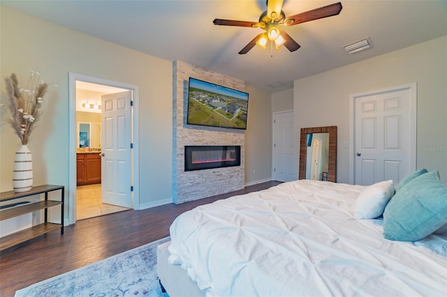 bedroom featuring a stone fireplace, hardwood / wood-style floors, connected bathroom, and ceiling fan