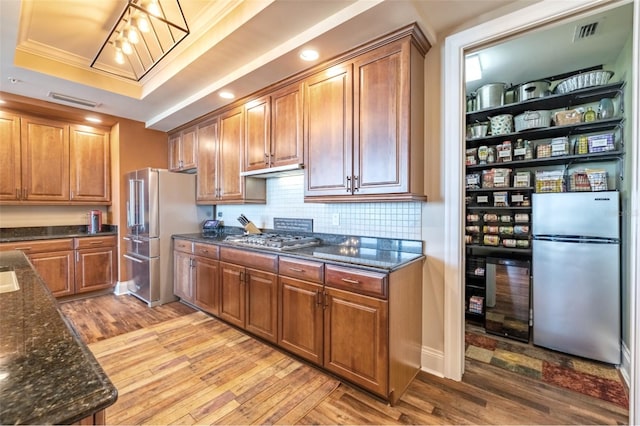 kitchen with a raised ceiling, dark stone countertops, stainless steel appliances, crown molding, and light wood-type flooring