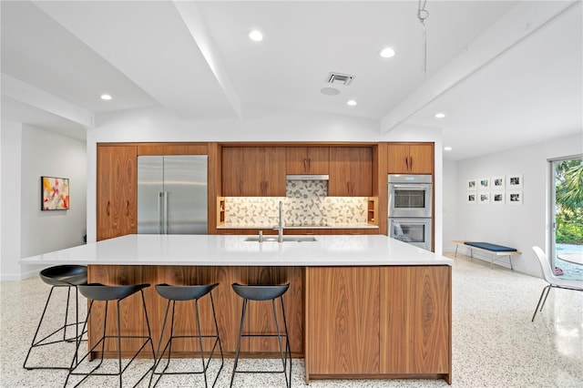 kitchen with sink, a center island with sink, stainless steel appliances, beam ceiling, and backsplash