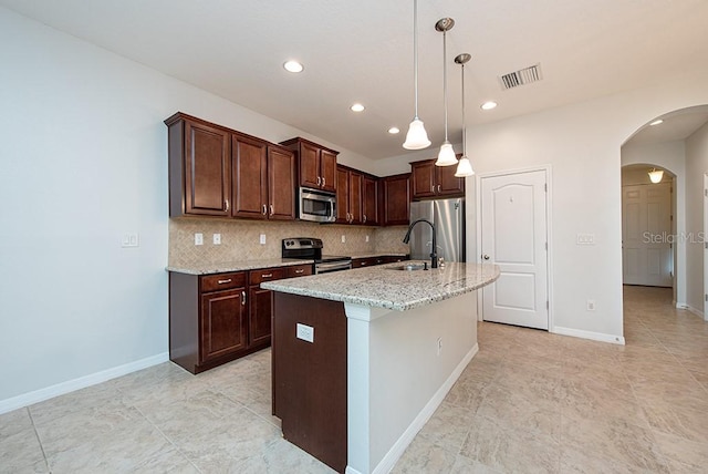 kitchen featuring appliances with stainless steel finishes, hanging light fixtures, backsplash, light stone counters, and a center island with sink