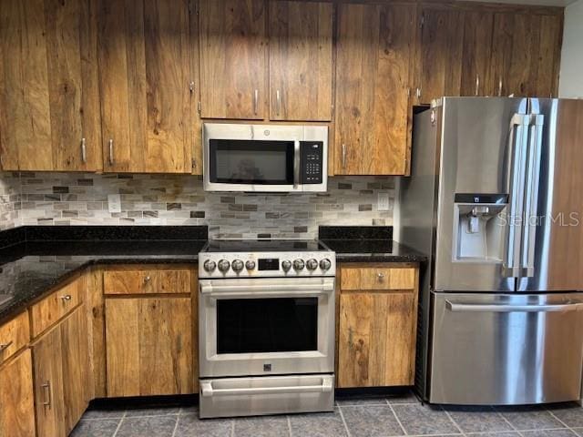 kitchen with brown cabinets, dark stone countertops, stainless steel appliances, and backsplash