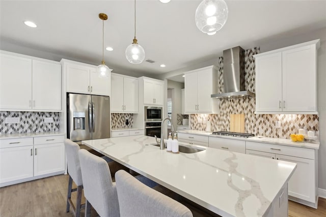 kitchen with an island with sink, a sink, appliances with stainless steel finishes, white cabinetry, and wall chimney range hood