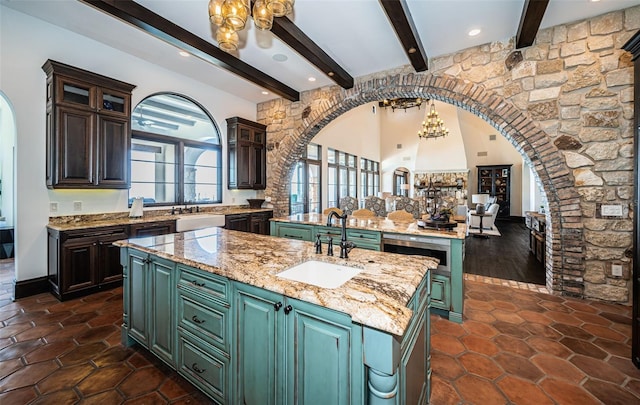 kitchen with arched walkways, glass insert cabinets, a sink, an island with sink, and light stone countertops