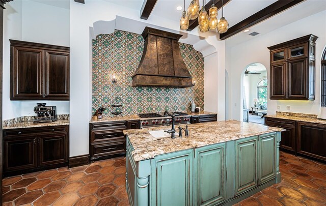 kitchen with arched walkways, hanging light fixtures, light stone countertops, a center island with sink, and custom range hood