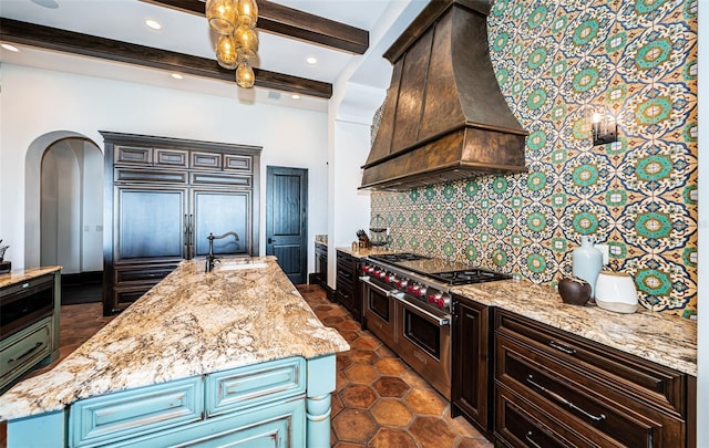 kitchen with range with two ovens, a sink, tasteful backsplash, an island with sink, and custom range hood