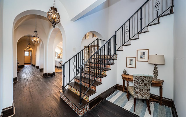 stairway featuring arched walkways, a notable chandelier, wood finished floors, high vaulted ceiling, and baseboards