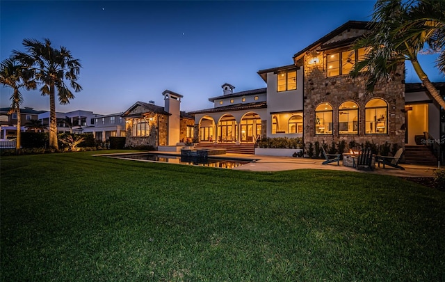 back of house at dusk with stone siding, a patio, and a lawn