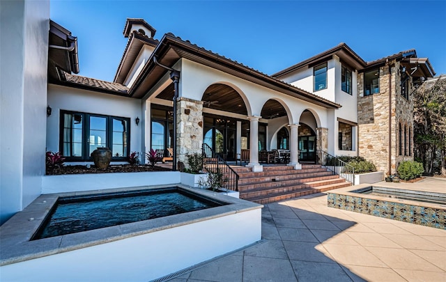 rear view of property featuring a tile roof, a patio, stucco siding, an in ground hot tub, and stone siding