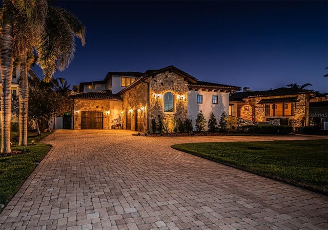 mediterranean / spanish home featuring an attached garage, stone siding, decorative driveway, and a yard