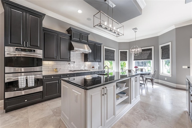 kitchen featuring appliances with stainless steel finishes, decorative light fixtures, an inviting chandelier, backsplash, and a kitchen island with sink