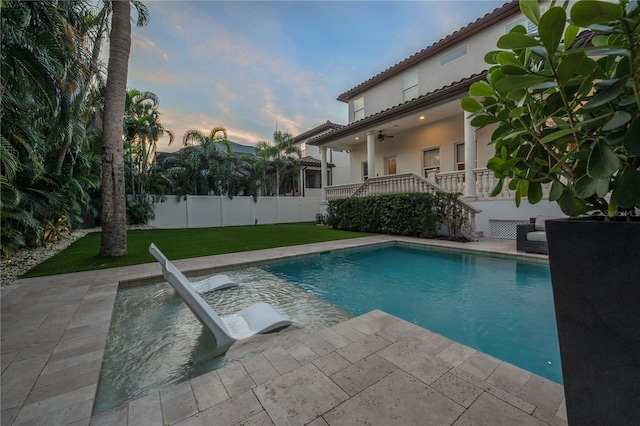 pool at dusk with ceiling fan, a patio area, and a yard