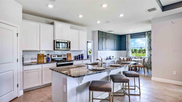 kitchen with white cabinets, appliances with stainless steel finishes, a kitchen island with sink, and dark stone countertops