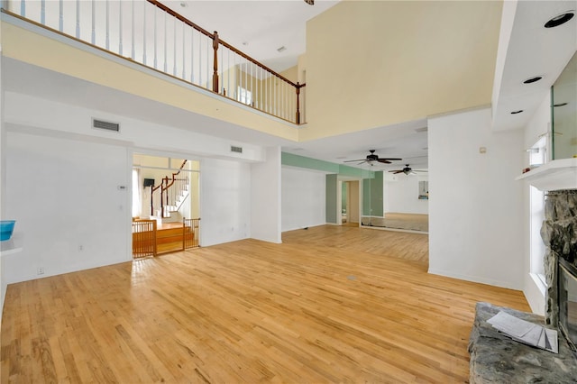 unfurnished living room featuring ceiling fan, a stone fireplace, light hardwood / wood-style flooring, and a high ceiling