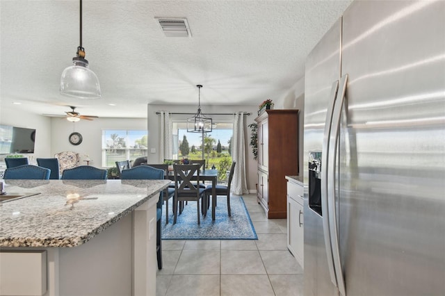 kitchen with light tile patterned floors, visible vents, stainless steel fridge with ice dispenser, hanging light fixtures, and light stone countertops