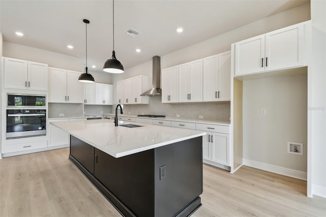 kitchen featuring wall chimney range hood, sink, a kitchen island with sink, white cabinetry, and stainless steel oven