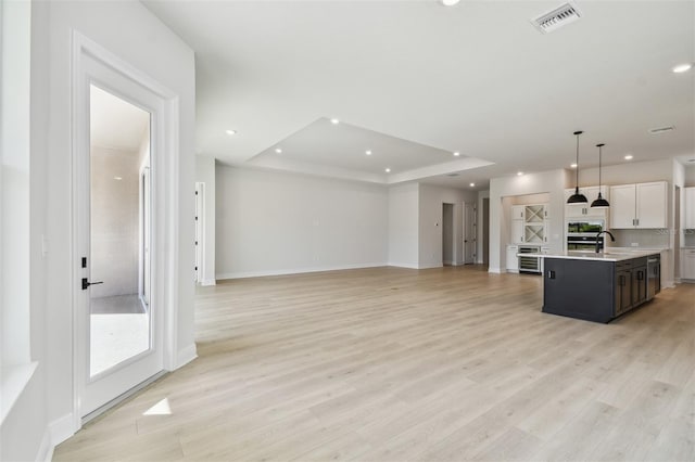 kitchen with white cabinetry, decorative light fixtures, a tray ceiling, an island with sink, and light hardwood / wood-style floors