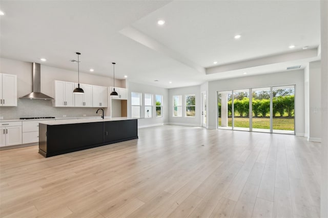 kitchen featuring wall chimney exhaust hood, black cooktop, white cabinetry, hanging light fixtures, and an island with sink