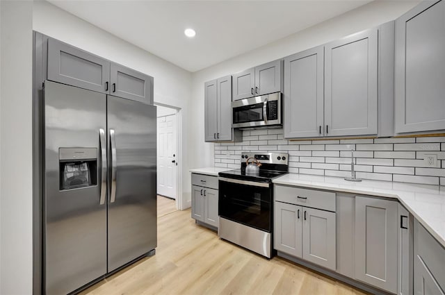 kitchen with gray cabinets, light hardwood / wood-style flooring, appliances with stainless steel finishes, backsplash, and light stone counters