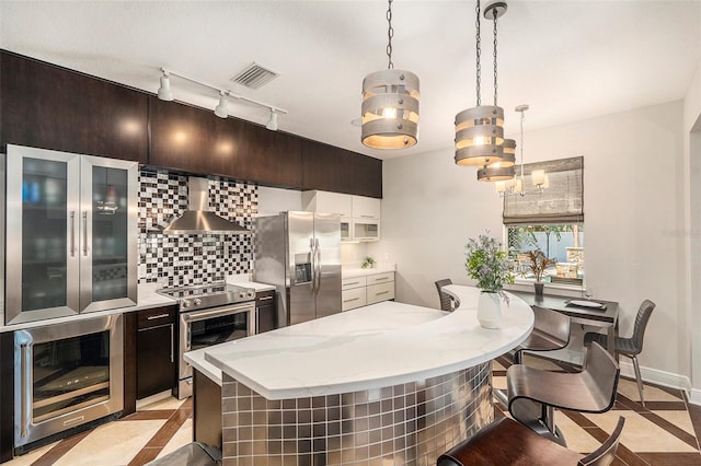 kitchen with appliances with stainless steel finishes, wall chimney exhaust hood, wine cooler, backsplash, and hanging light fixtures