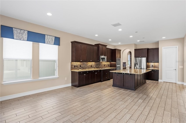 kitchen featuring sink, appliances with stainless steel finishes, a kitchen island with sink, decorative backsplash, and light wood-type flooring