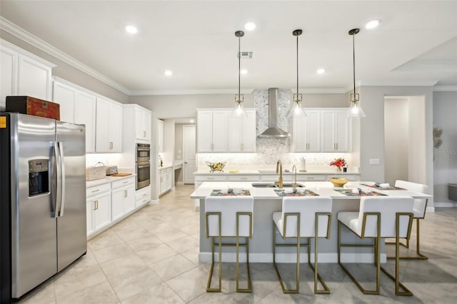 kitchen with wall chimney range hood, appliances with stainless steel finishes, a kitchen island with sink, white cabinetry, and decorative light fixtures