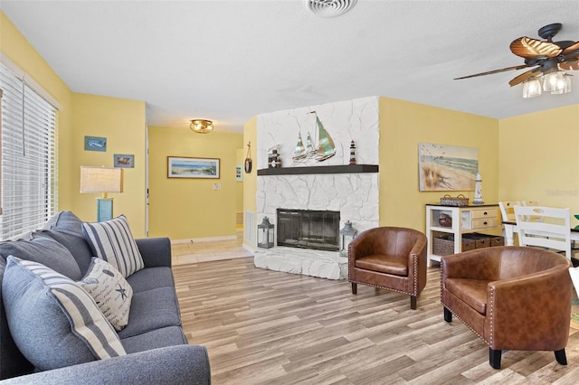 living room featuring ceiling fan, a stone fireplace, a textured ceiling, and light hardwood / wood-style flooring