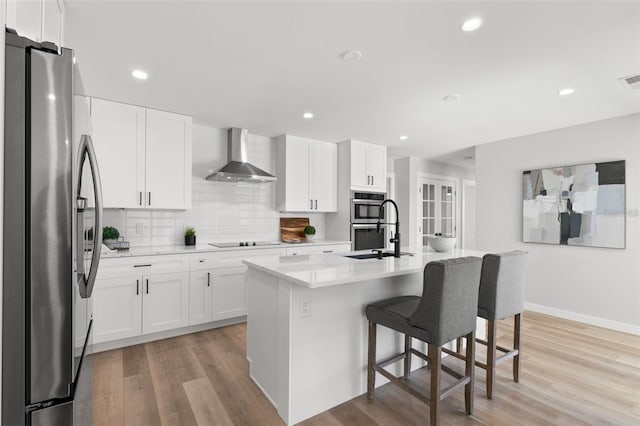 kitchen with appliances with stainless steel finishes, white cabinetry, a breakfast bar area, an island with sink, and wall chimney exhaust hood