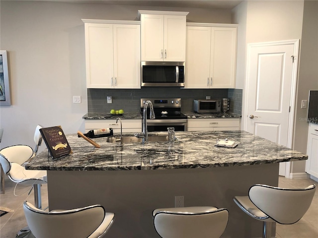 kitchen featuring white cabinetry, a center island with sink, stainless steel appliances, and dark stone countertops