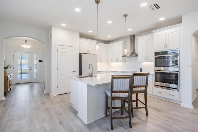 kitchen with hanging light fixtures, a kitchen island with sink, white cabinets, and wall chimney exhaust hood