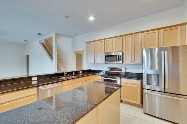 kitchen with light tile patterned floors, dark stone counters, appliances with stainless steel finishes, light brown cabinetry, and a sink