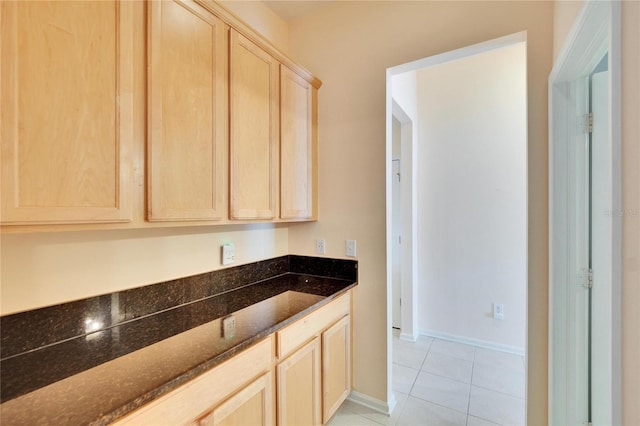 kitchen with light tile patterned floors, baseboards, dark stone countertops, and light brown cabinetry