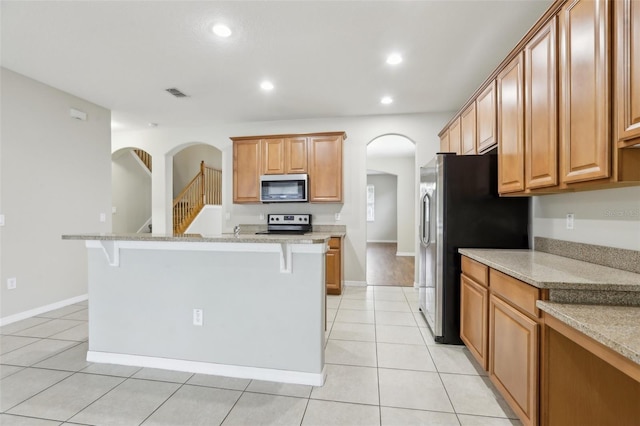 kitchen with appliances with stainless steel finishes, an island with sink, a breakfast bar area, light tile patterned floors, and light stone counters