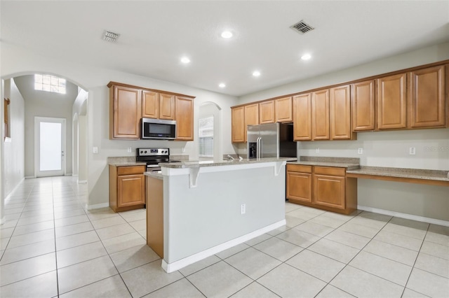 kitchen featuring appliances with stainless steel finishes, a breakfast bar, an island with sink, light tile patterned floors, and light stone counters