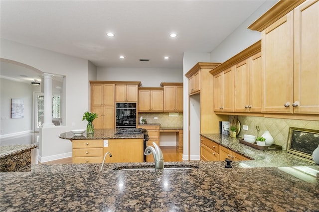 kitchen featuring ornate columns, light brown cabinetry, double oven, dark stone countertops, and decorative backsplash