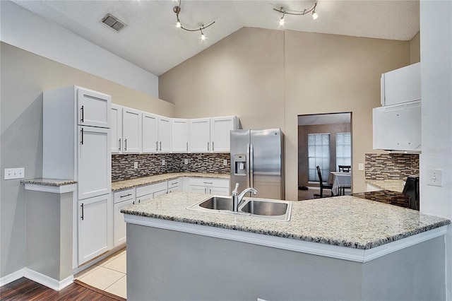 kitchen with high vaulted ceiling, white cabinetry, sink, stainless steel fridge, and light stone countertops