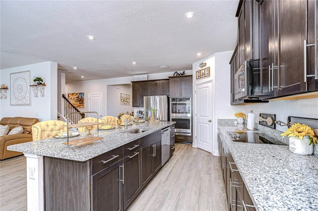 kitchen with appliances with stainless steel finishes, sink, a kitchen island with sink, light stone counters, and light hardwood / wood-style flooring