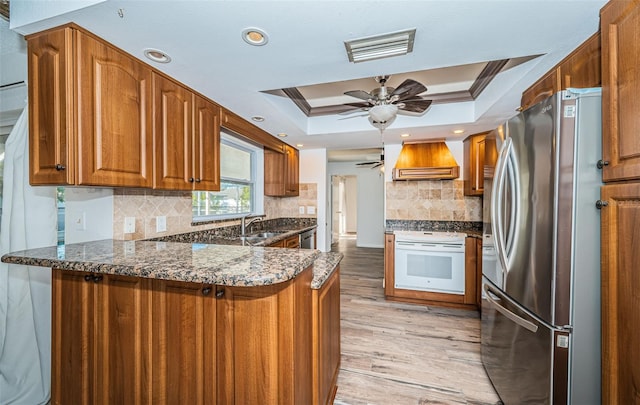 kitchen with premium range hood, appliances with stainless steel finishes, dark stone countertops, kitchen peninsula, and a raised ceiling