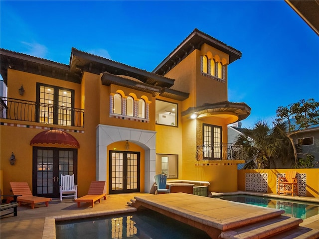 back house at dusk featuring french doors, a balcony, a swimming pool with hot tub, and a patio area