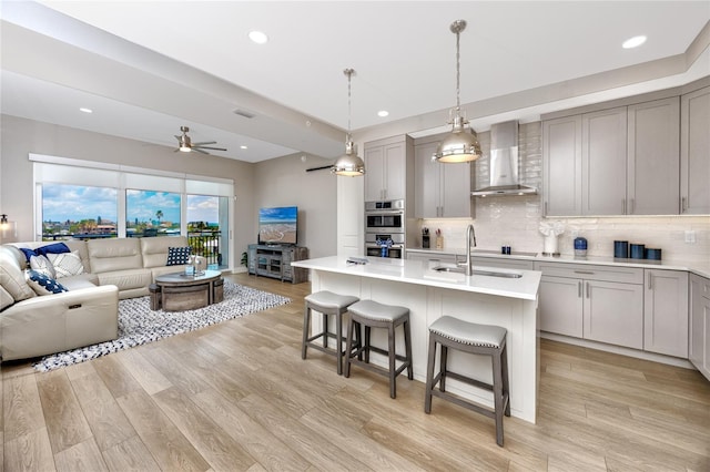 kitchen with gray cabinets, an island with sink, sink, and wall chimney range hood