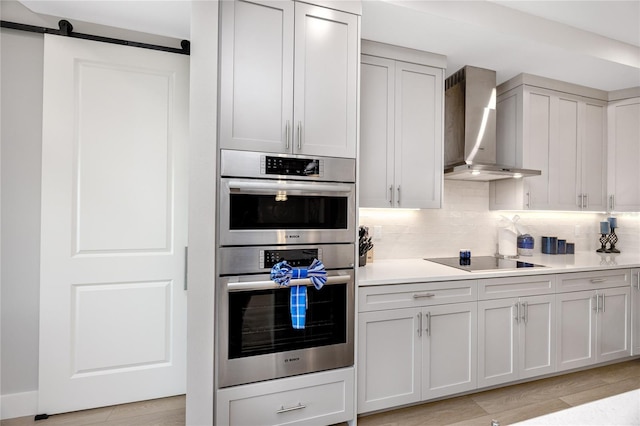 kitchen featuring wall chimney range hood, tasteful backsplash, black electric cooktop, a barn door, and stainless steel double oven