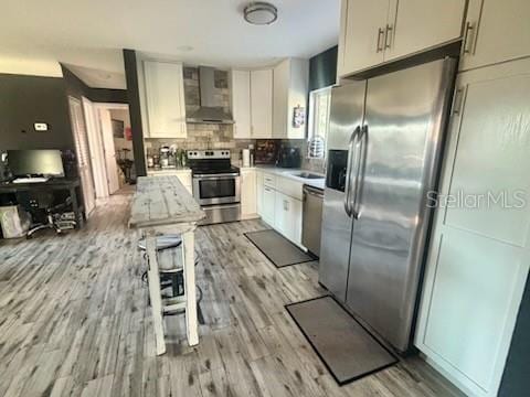 kitchen featuring wall chimney exhaust hood, white cabinetry, appliances with stainless steel finishes, light hardwood / wood-style floors, and decorative backsplash