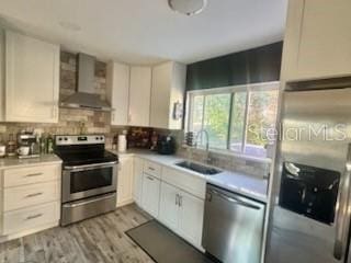 kitchen with wall chimney range hood, sink, light hardwood / wood-style flooring, appliances with stainless steel finishes, and white cabinets