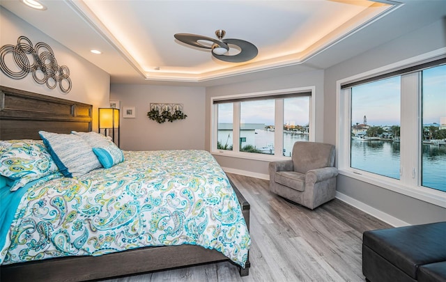 bedroom featuring a raised ceiling, wood-type flooring, a water view, and ceiling fan