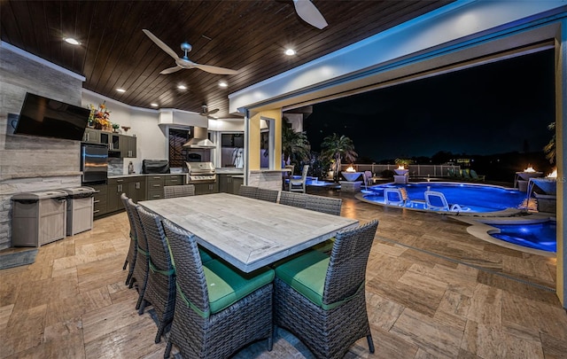 patio at twilight with area for grilling, a bar, ceiling fan, a grill, and a fenced in pool