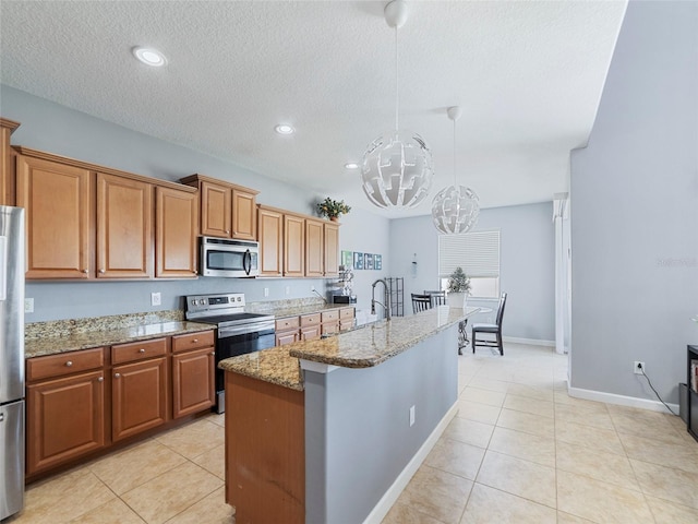 kitchen with light tile patterned flooring, hanging light fixtures, appliances with stainless steel finishes, an island with sink, and light stone countertops