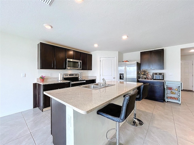 kitchen featuring appliances with stainless steel finishes, an island with sink, sink, light tile patterned floors, and dark brown cabinets