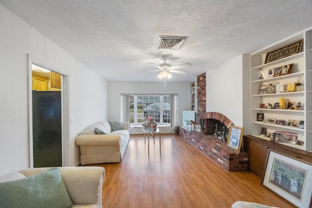 living room with built in shelves, a brick fireplace, a textured ceiling, hardwood / wood-style flooring, and ceiling fan