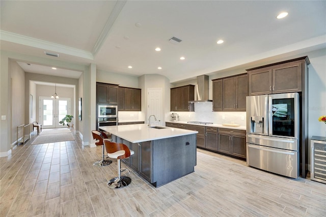 kitchen with wine cooler, wall chimney exhaust hood, sink, a center island with sink, and appliances with stainless steel finishes