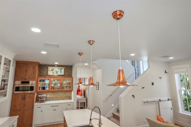 kitchen with stainless steel oven, white cabinets, brown cabinetry, glass insert cabinets, and pendant lighting