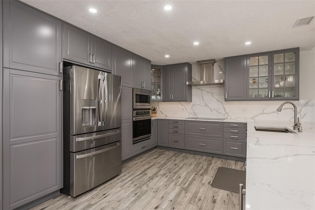 kitchen with sink, gray cabinetry, stainless steel appliances, light stone countertops, and wall chimney range hood